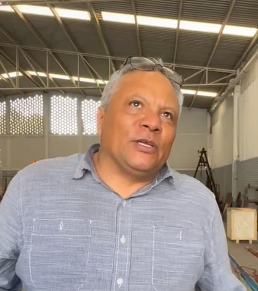 Una persona con cabello corto y gafas en la cabeza viste una camisa de color claro y está parada en el interior de un edificio con techo alto y equipo industrial.